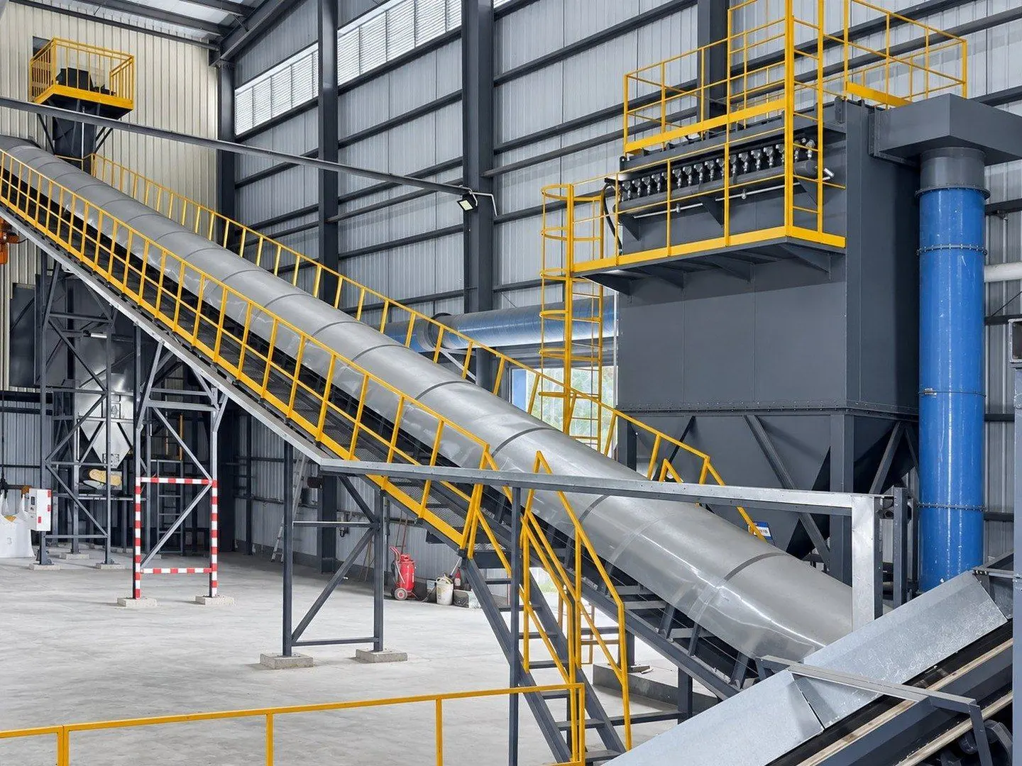 Long enclosed inclined belt conveyor with a walkway and yellow handrails, rising across a workshop