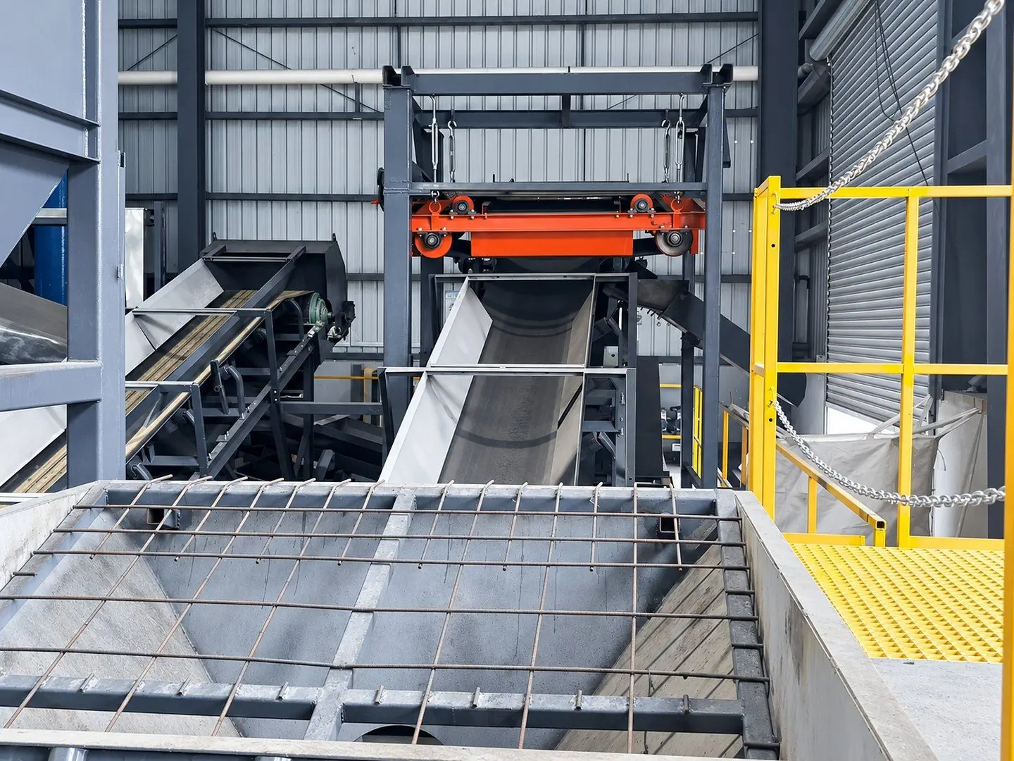 Grated hopper in front of a belt conveyor running under a red overband magnet